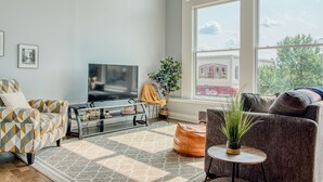 Ein Wohnzimmer mit großem Fenster, Flachbildfernseher, gemusterten Sesseln, Couchtisch und einer Topfpflanze.