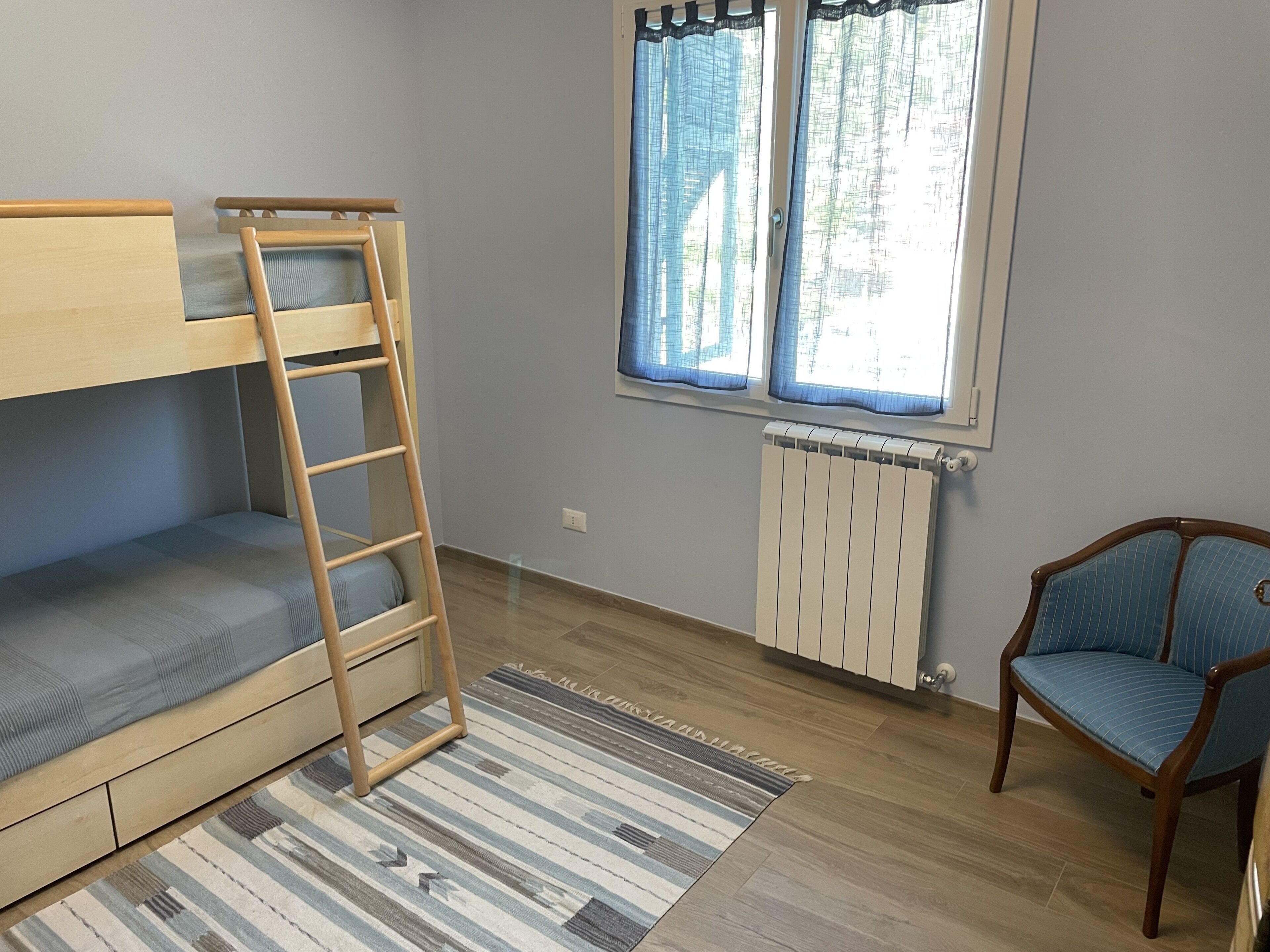 Une chambre avec un lit superposé, une échelle, une chaise, une fenêtre avec des rideaux et un radiateur.