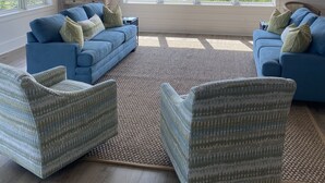 Un salón amplio con dos sofás azules, un espejo redondo y un ventilador de techo. Amplias ventanas ofrecen vistas al exterior.