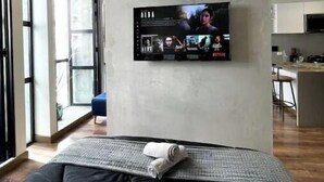Moderni huone, jossa on suuri seinälle asennettu taulutelevisio, pehmustetulla päällisellä varustettu sänky ja näkymä keittiötilaan lasiovien läpi.