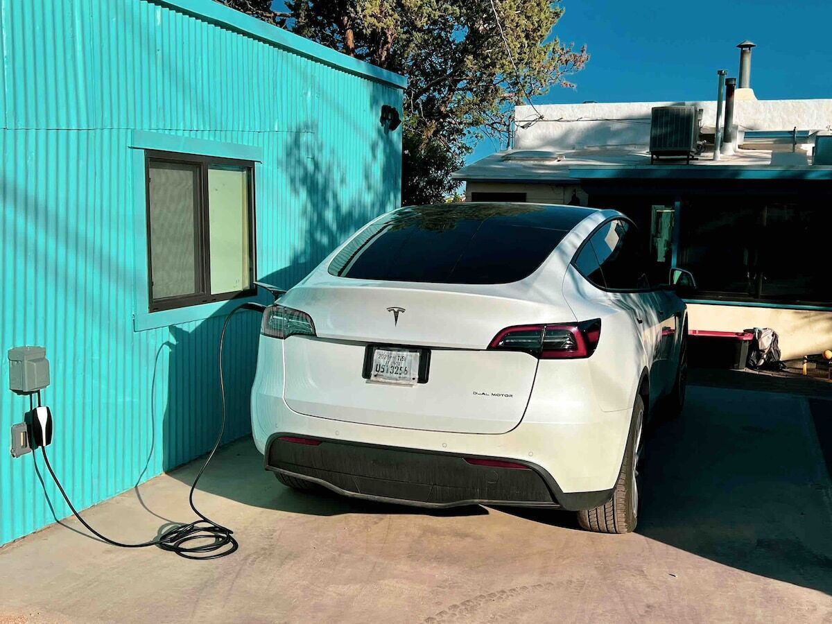 A white Tesla Model 3 parked in a garage with a turquoise building and a clear blue sky in the background.
