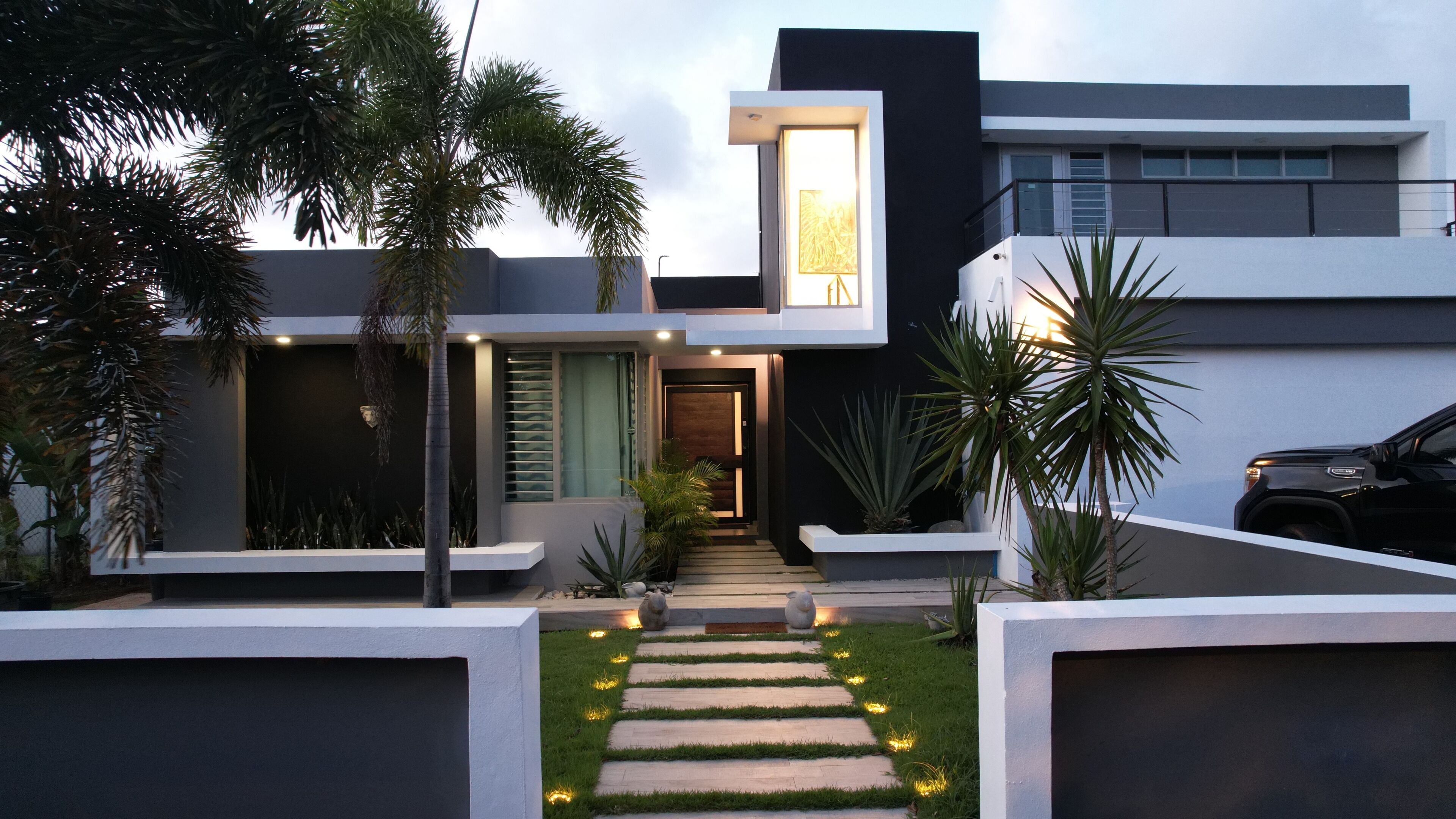 Una casa moderna con diseño minimalista, que presenta un camino central que lleva a la entrada, rodeada de plantas en macetas y un césped bien cuidado.