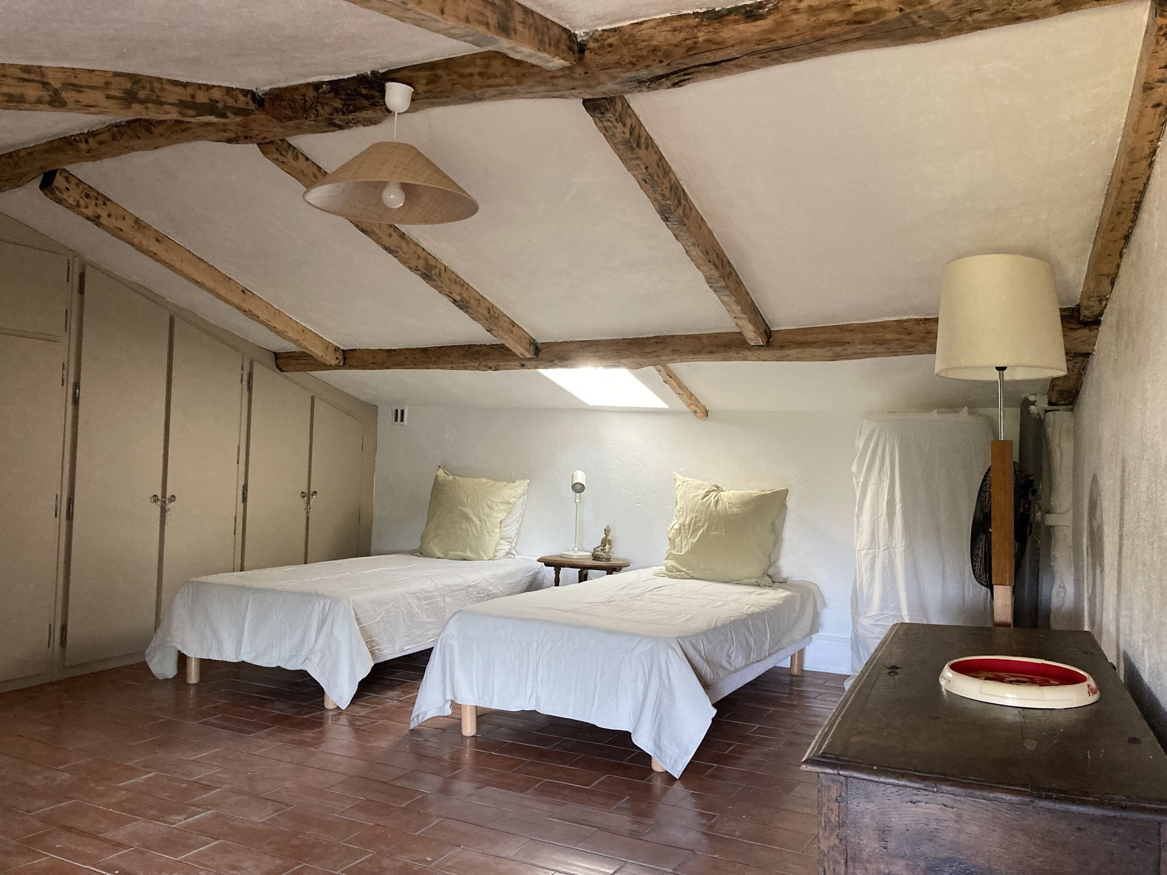 Une chambre avec deux lits simples, des poutres en bois au plafond, une petite table avec une lampe et un miroir à cadre rouge.