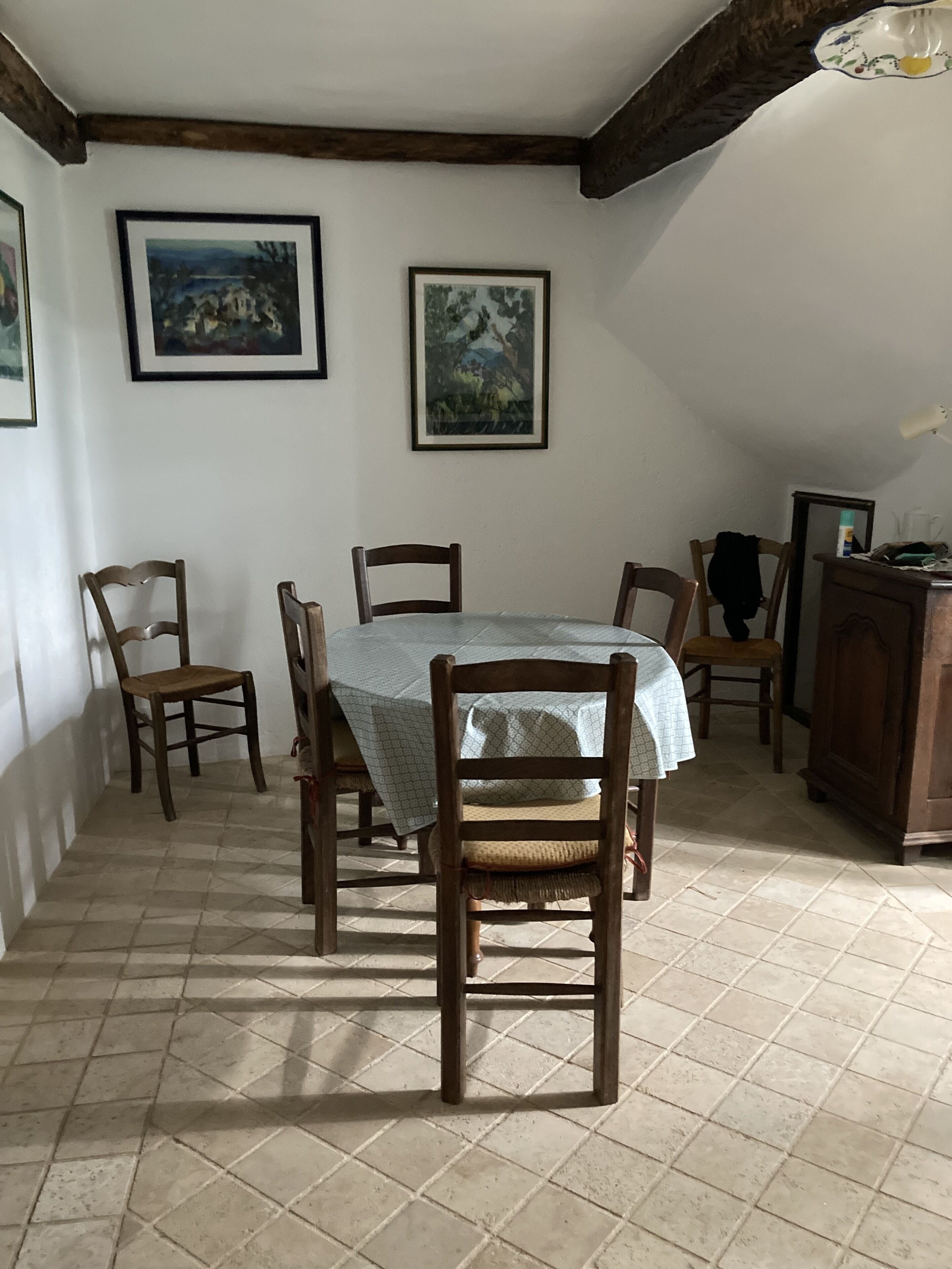 Ein Essbereich mit Tisch, gedeckt für vier Personen, umgeben von Holzstühlen. An der Wand über dem Tisch hängen zwei gerahmte Gemälde. Der Raum hat einen gefliesten Boden und einen Holzbalken an der Decke.