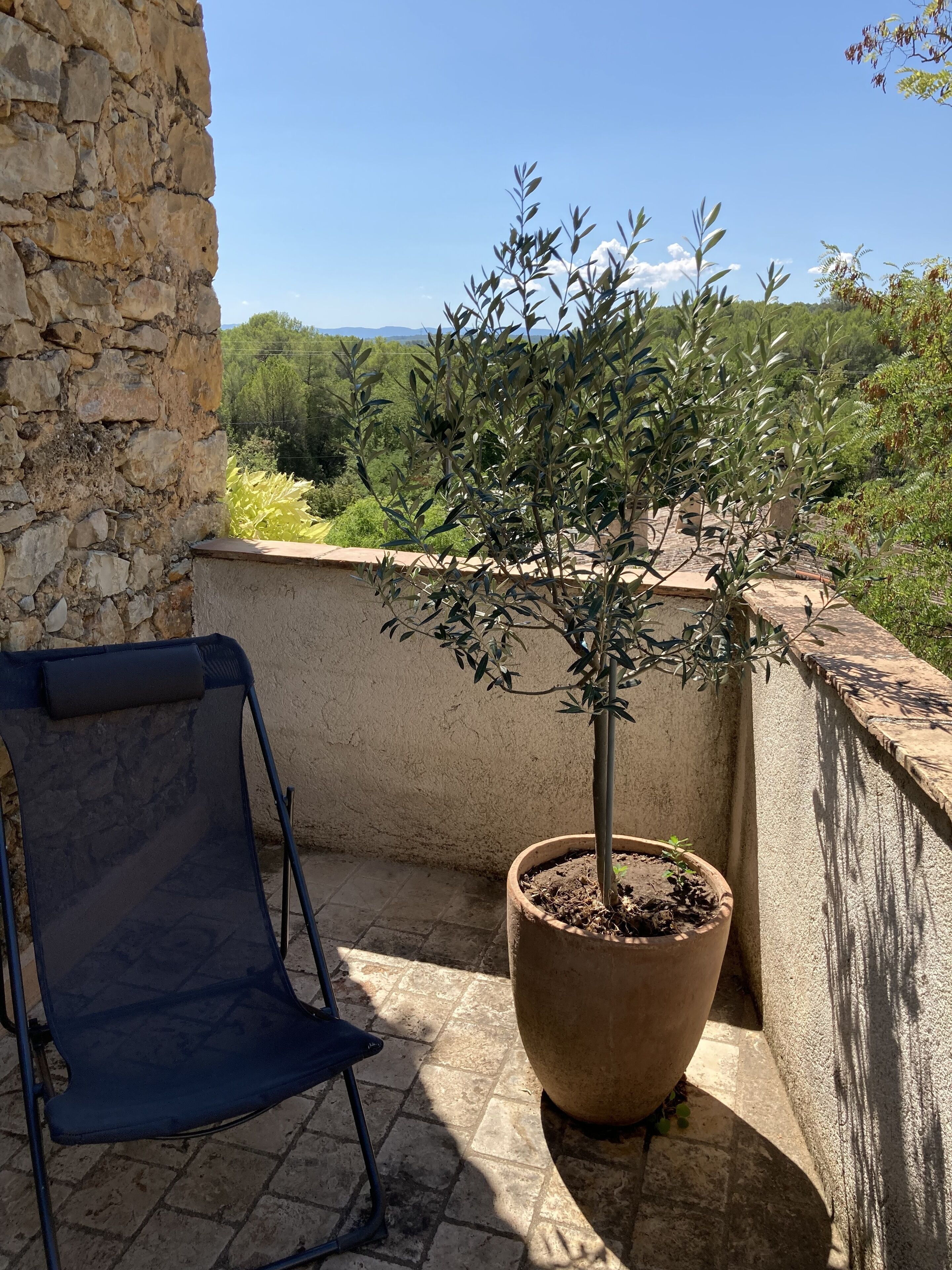 Un balcon orné d’un olivier en pot, d’une chaise pliante bleue et d’un mur en pierre.
