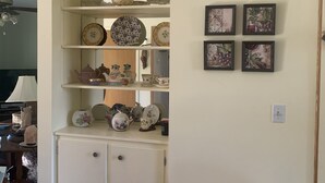 Una habitación con un aparador blanco con varios estantes que contienen diversos objetos decorativos y platos. Hay cuatro cuadros enmarcados en la pared a la derecha.