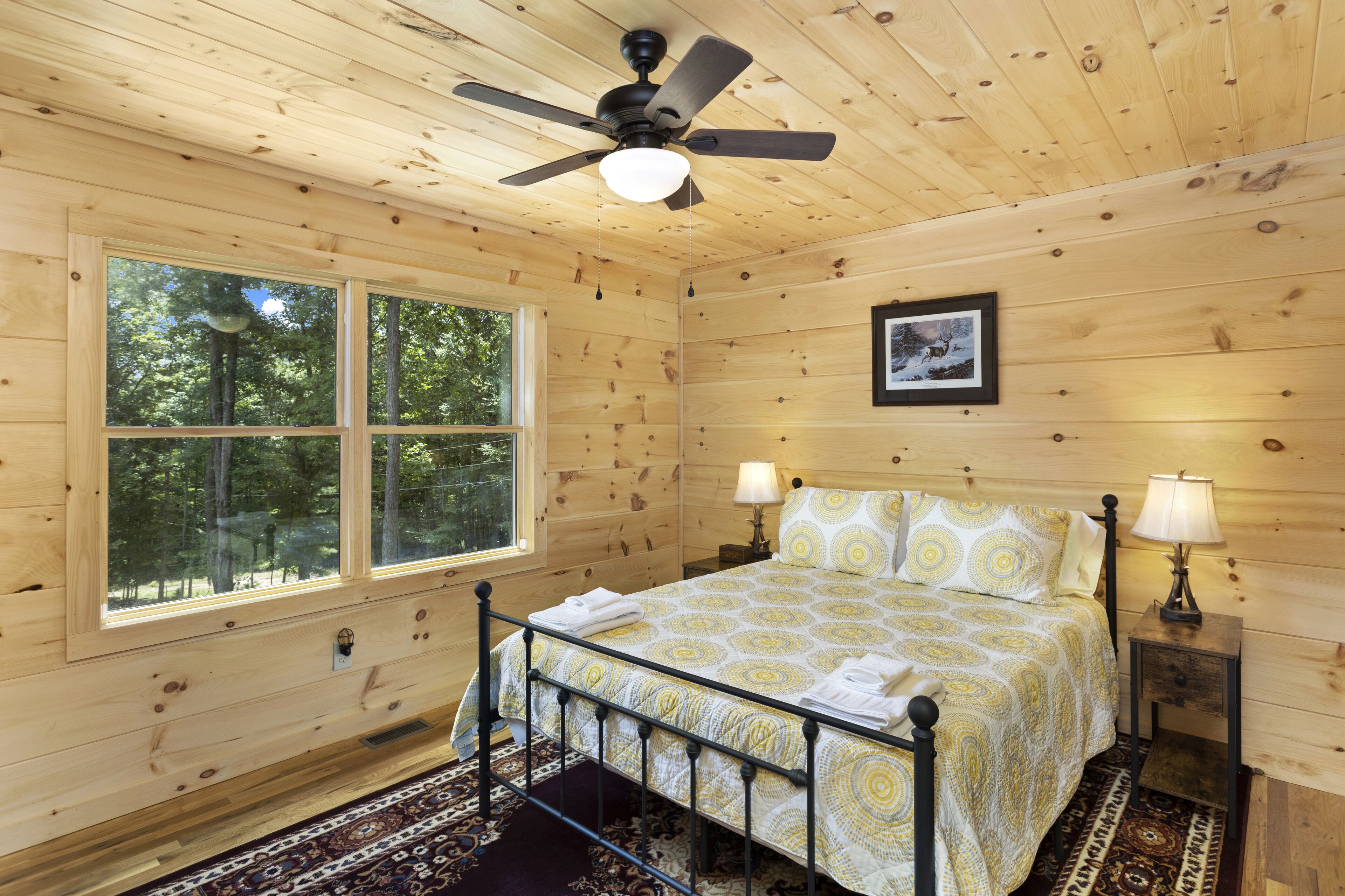 A wooden cabin bedroom with a bed, two bedside lamps, a ceiling fan, and a window with a view of trees.