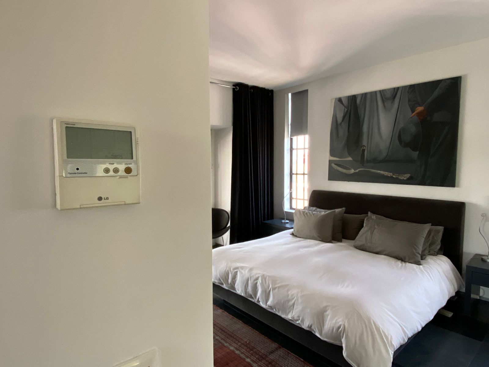 A hotel room with a bed, a nightstand, a wall-mounted thermostat, and a large abstract painting.
