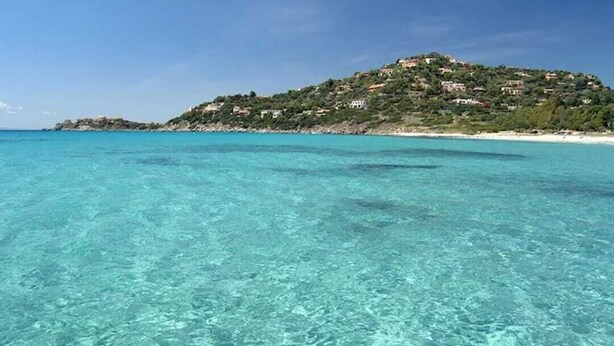 Cena costeira com águas turquesa cristalinas, uma praia arenosa e uma colina com edifícios.