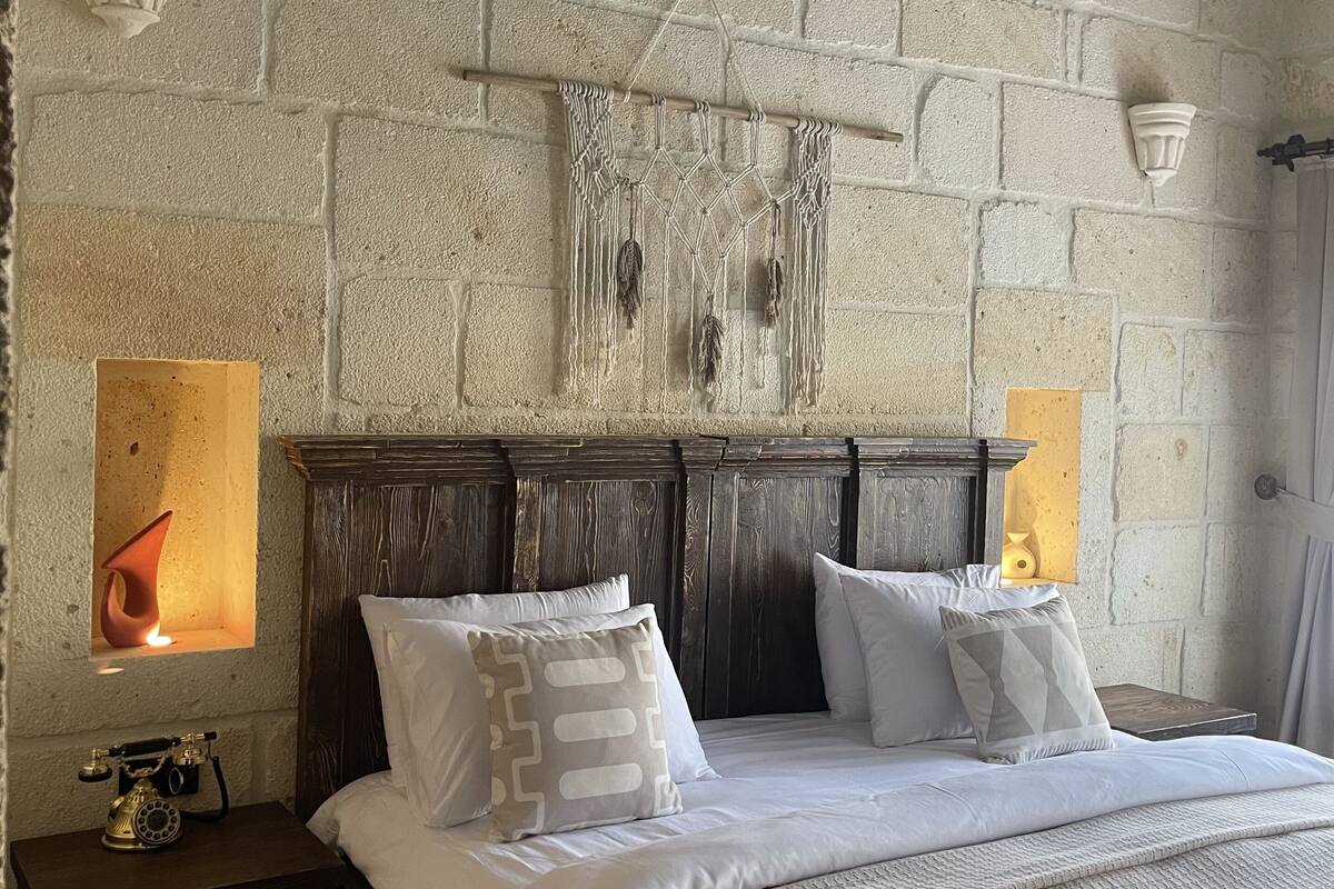 A bedroom with a brick wall, wooden headboard, white bedding, and a macrame wall hanging.