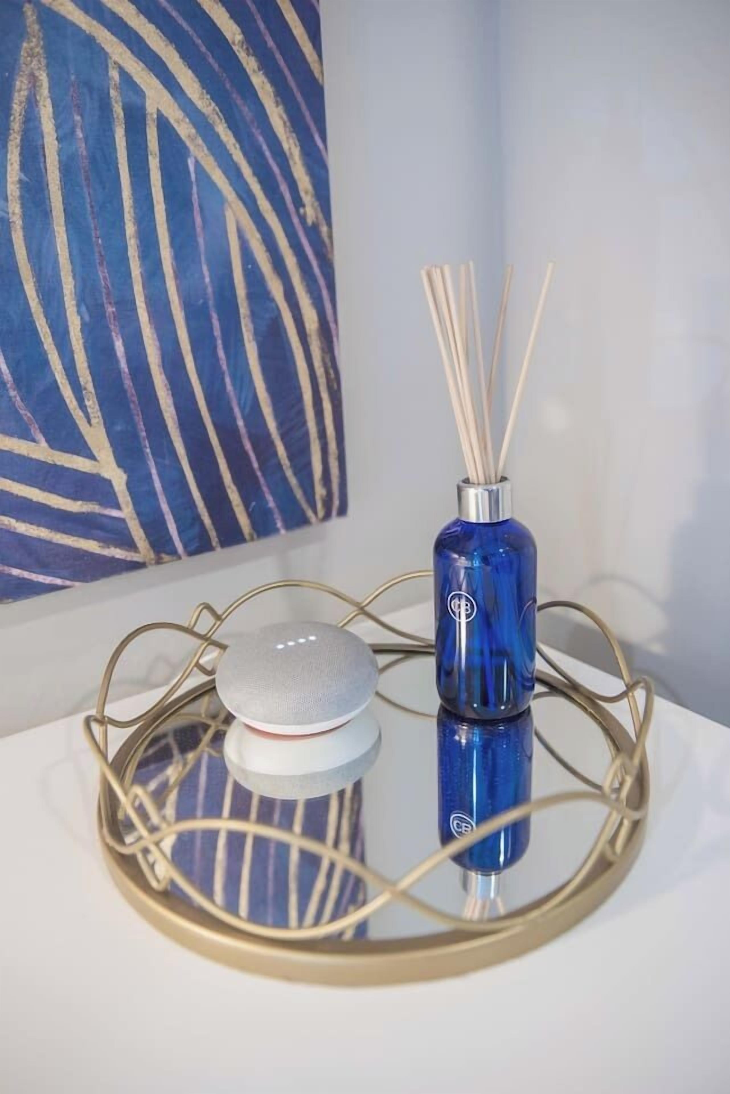 Un diffuseur bleu avec des tiges en roseau, posé sur un plateau réfléchissant, un objet blanc et un tableau abstrait bleu accroché au mur.