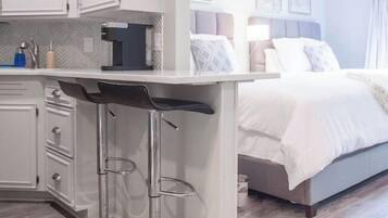 A modern hotel room with a kitchenette, a bed with white linens, and a small seating area with stools.