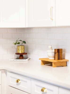 Una cocina con armarios blancos, salpicadero de azulejos blancos, tabla de cortar de madera y una maceta dorada decorativa sobre un pedestal blanco.