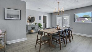 A dining area with a wooden table and chairs, a chandelier, and a view of the outdoors through a window.