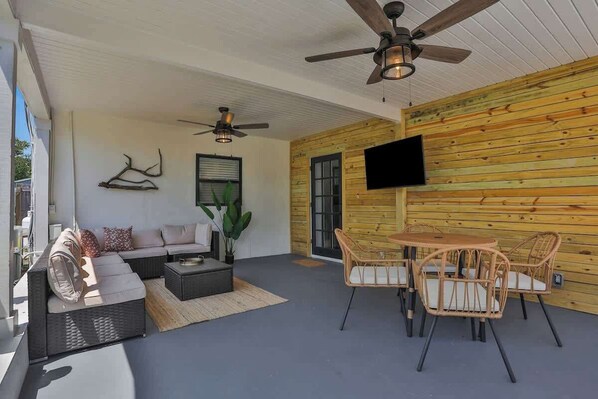 A covered outdoor area with a sofa, chairs, a table, ceiling fans, and a mounted TV.