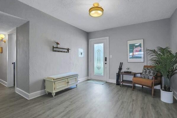 A room with a light-colored wall, a wooden floor, a bench, a chair, a potted plant, and a door with a frosted glass panel.