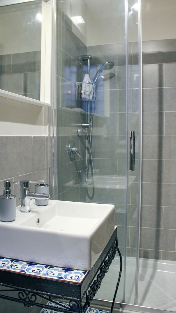 Un baño moderno con cabina de ducha de vidrio, un lavabo blanco con un borde de azulejos decorativos y un cabezal de ducha de mano.