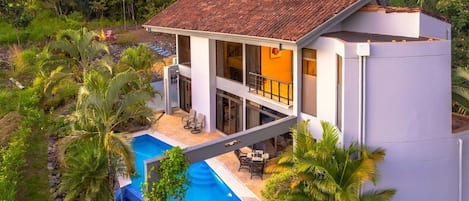 A modern two-story house with a large balcony, a swimming pool, and lush tropical vegetation.