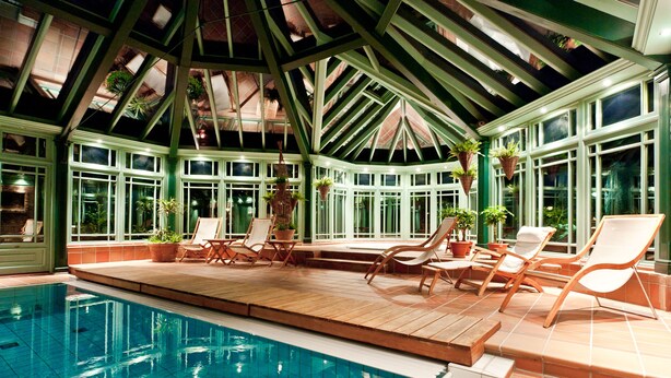 An indoor swimming pool with wooden deck and lounge chairs, surrounded by large windows and potted plants. - Engø Gård (Færder)
