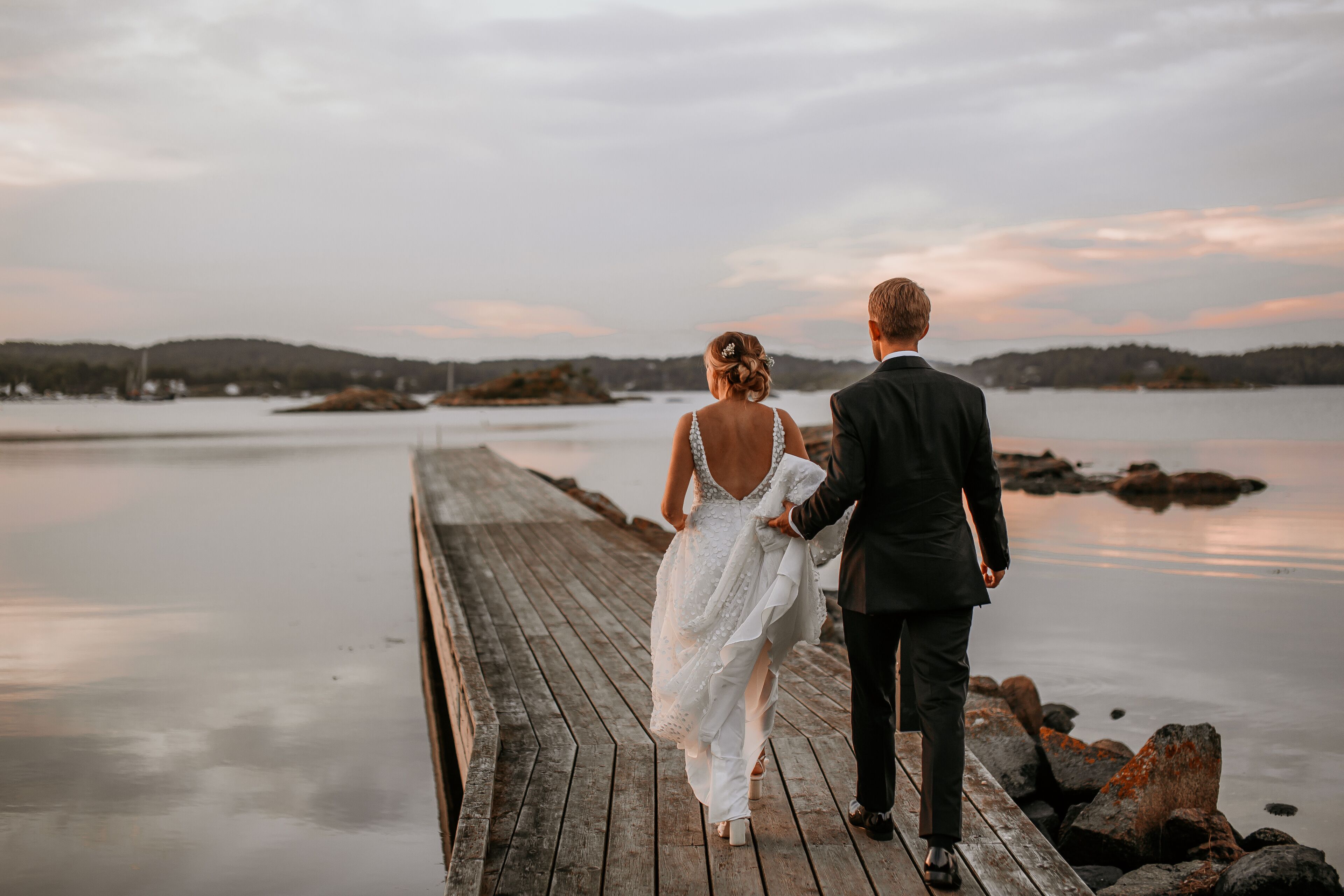 A couple walking on a wooden pier with a calm lake and distant hills in the background.