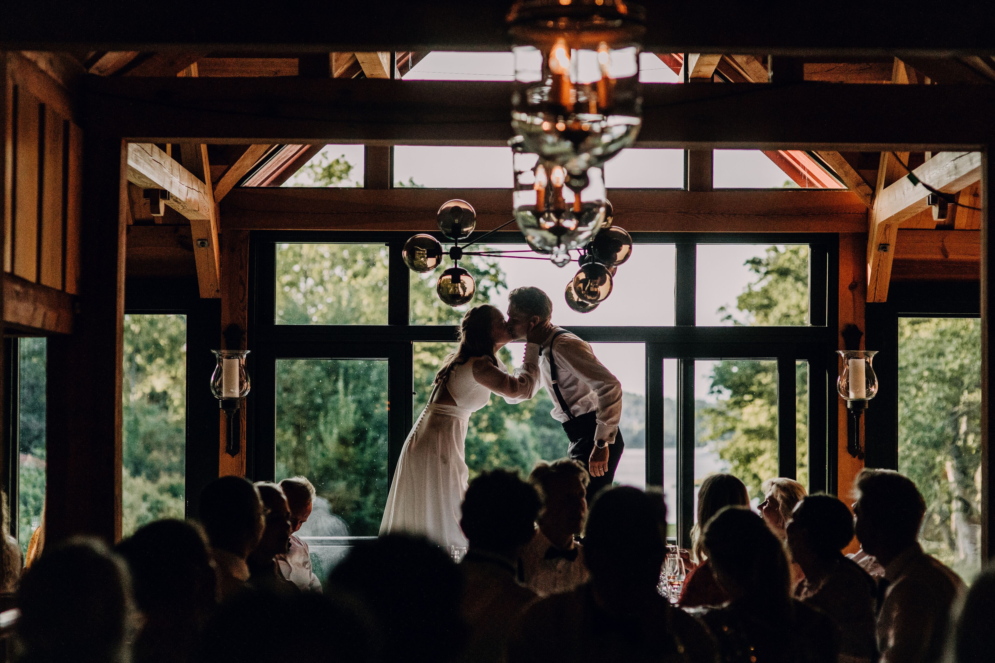 A couple kissing in a rustic wooden room with large windows and hanging light fixtures.
