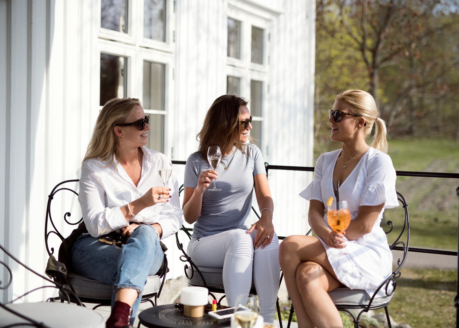 Trois femmes sont assises sur des chaises en métal à l’extérieur, autour d’une table où sont posées des boissons, devant un bâtiment blanc doté de grandes fenêtres.