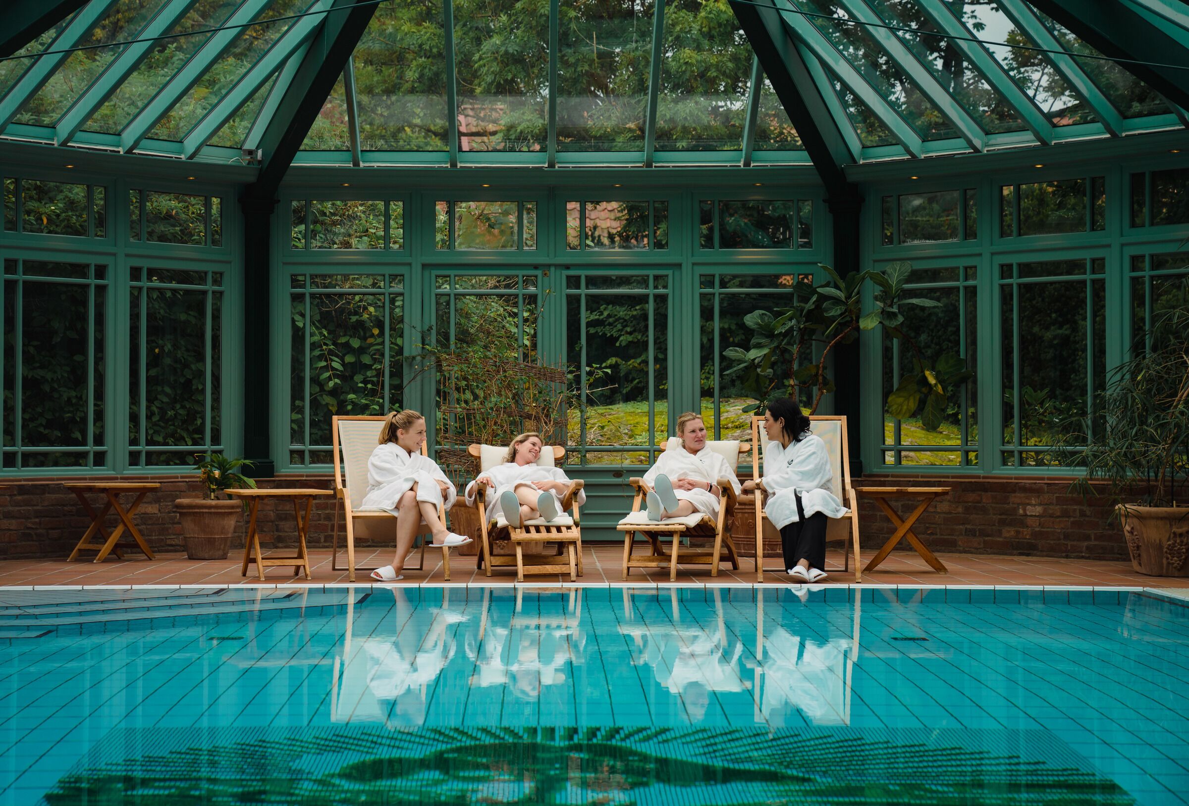 Zona de piscina con una estructura de vidrio, sillones de madera y una piscina cristalina.