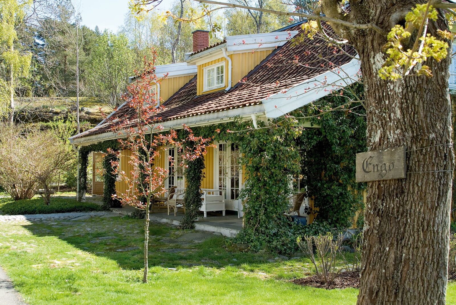 Een traditioneel huis met een overdekte veranda, een bordje met de tekst 'Enzo' en een goed onderhouden tuin.