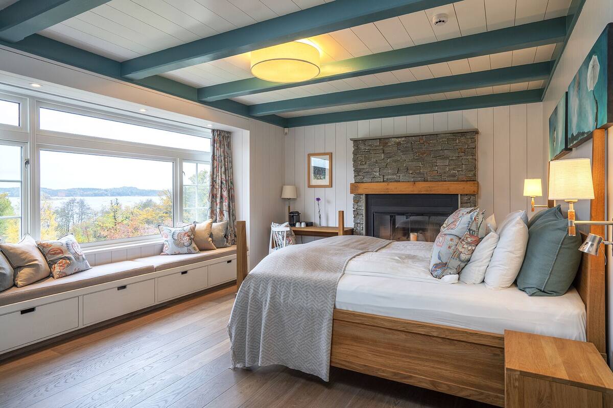 A bedroom with a large bed, a stone fireplace, a window seat with cushions, and a view of the outdoors.