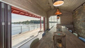 A modern interior with a long wooden table, pendant lighting, and a view of a river. - Engø Gård (Færder)