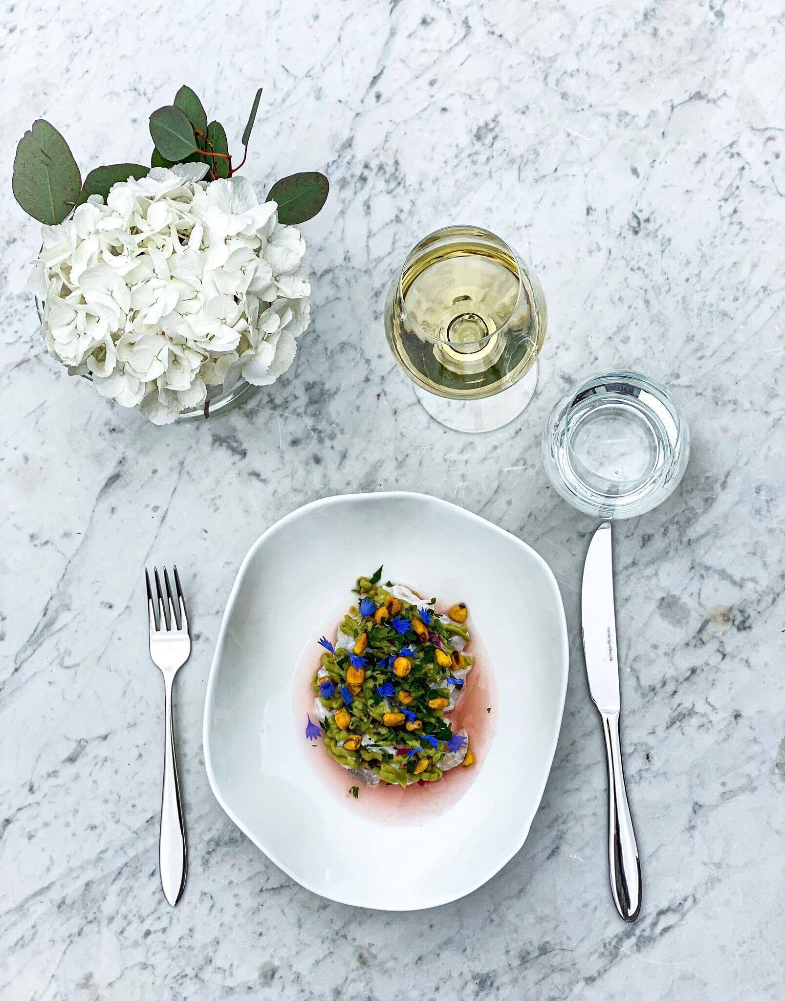 Une assiette blanche contenant un plat, une fourchette, un couteau, un verre de vin blanc et un petit verre d’eau, le tout posé sur une surface en marbre.