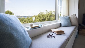 A cozy reading nook with a view of a lake and trees, featuring a cushioned bench, books, and glasses. - Engø Gård (Færder)