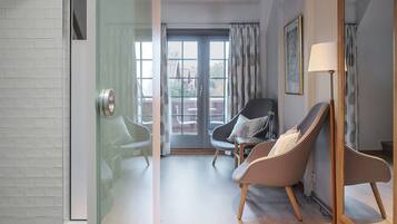 A modern living room with a glass partition, two armchairs, a floor lamp, and a view through a glass door to an outdoor area.