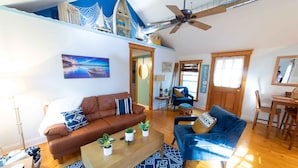 A spacious living area with a brown leather sofa, a blue armchair, and a wooden coffee table with plants. A ceiling fan and a nautical-themed shelf are visible. - The charming Glenn Cottage! Walk to Lake Michigan! (Fennville)