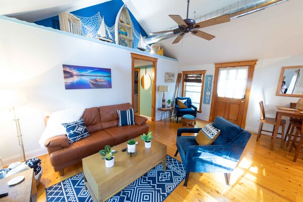 A spacious living area with a brown sofa, blue armchair, and a coffee table with plants. A ceiling fan and a painting of a boat are visible.