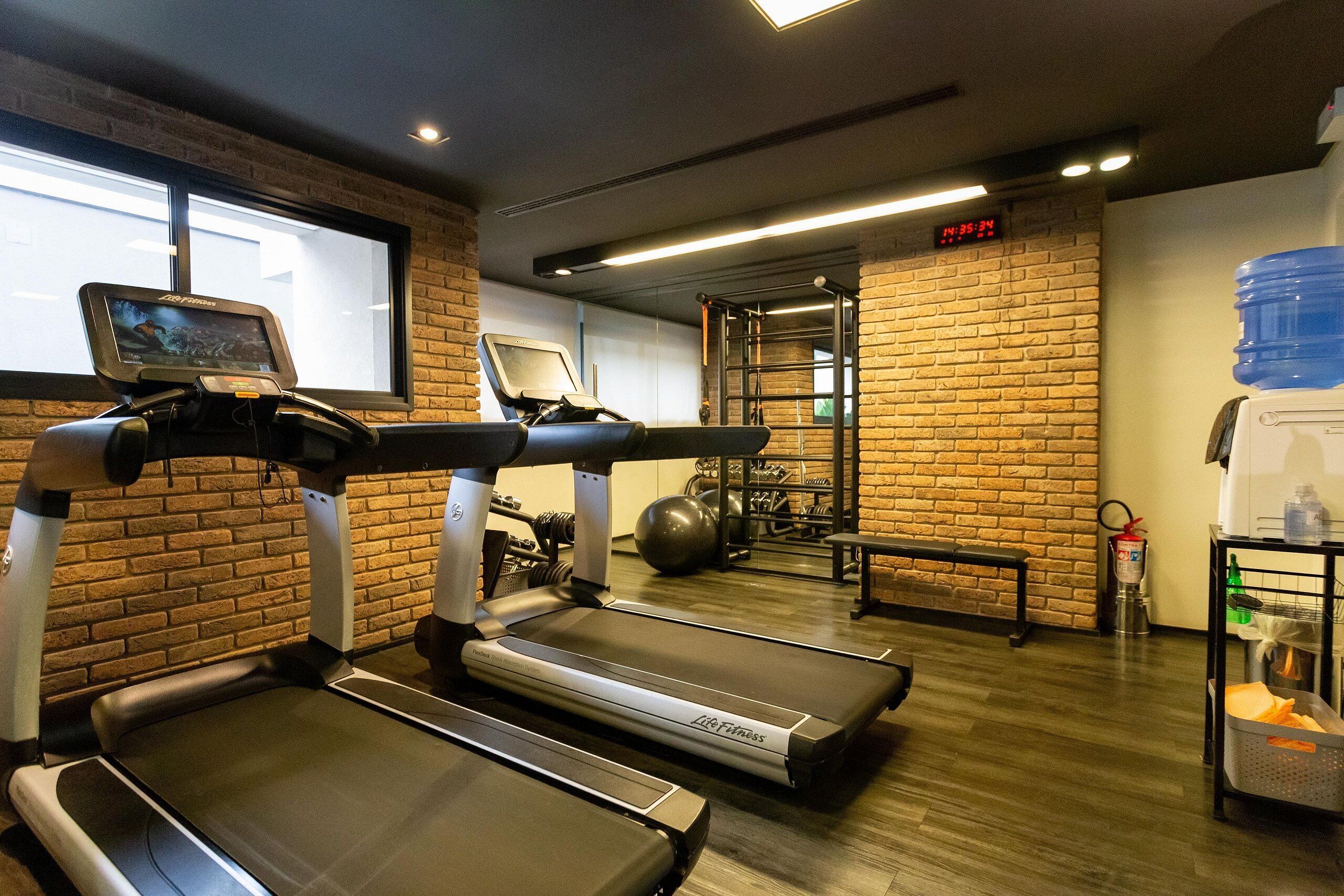 Un gimnasio con cintas de correr, una pantalla digital, una pared de ladrillo, un dispensador de agua y un espejo.