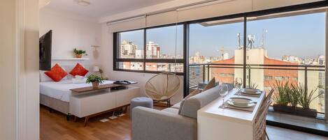 A modern apartment with a large window, a bed, a sofa, a dining table, and a TV.