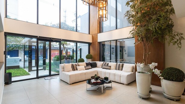 A spacious living area with a large sectional sofa, a coffee table, and potted plants. - Condomínio Edifício Living Design (São Paulo)