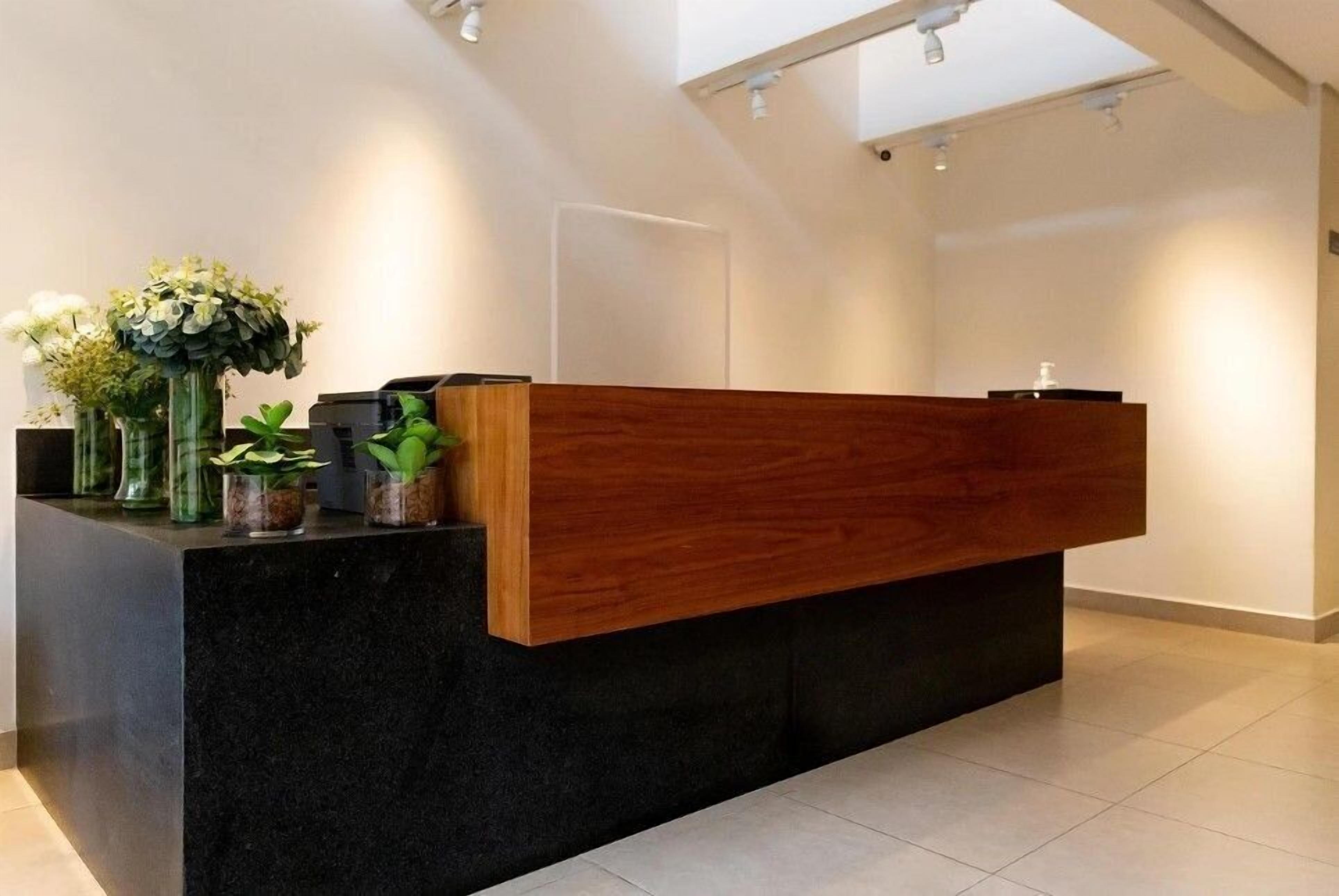 Una mesa de recepción con frente de madera y encimera negra, flanqueada por plantas en macetas.
