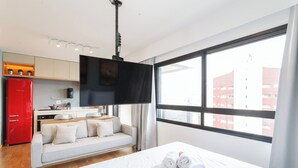 A modern hotel room with a bed, a sofa, a kitchen area, and a large window offering a city view. - Condomínio Edifício Living Design (São Paulo)