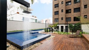 A rooftop pool with a waterfall feature, surrounded by wooden decking and a seating area with chairs. - Condomínio Edifício Living Design (São Paulo)
