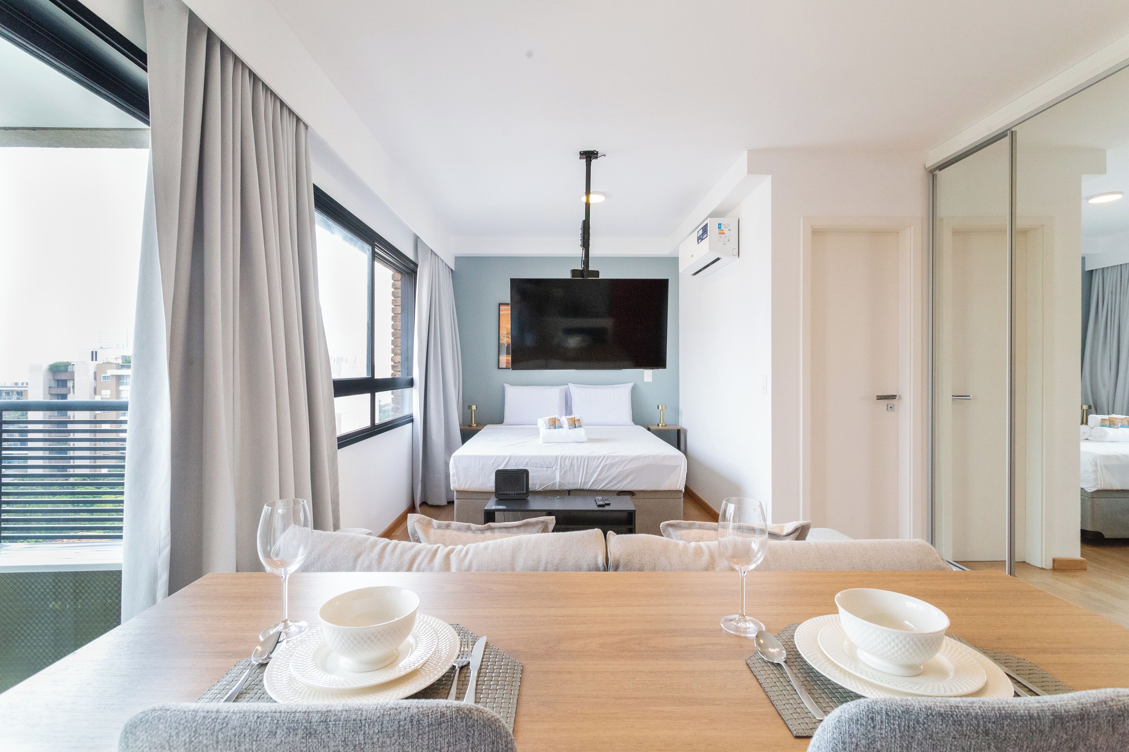 Habitación de hotel con cama, televisor montado en la pared, mesa de comedor con sillas y vista a la ciudad a través de ventanas amplias.