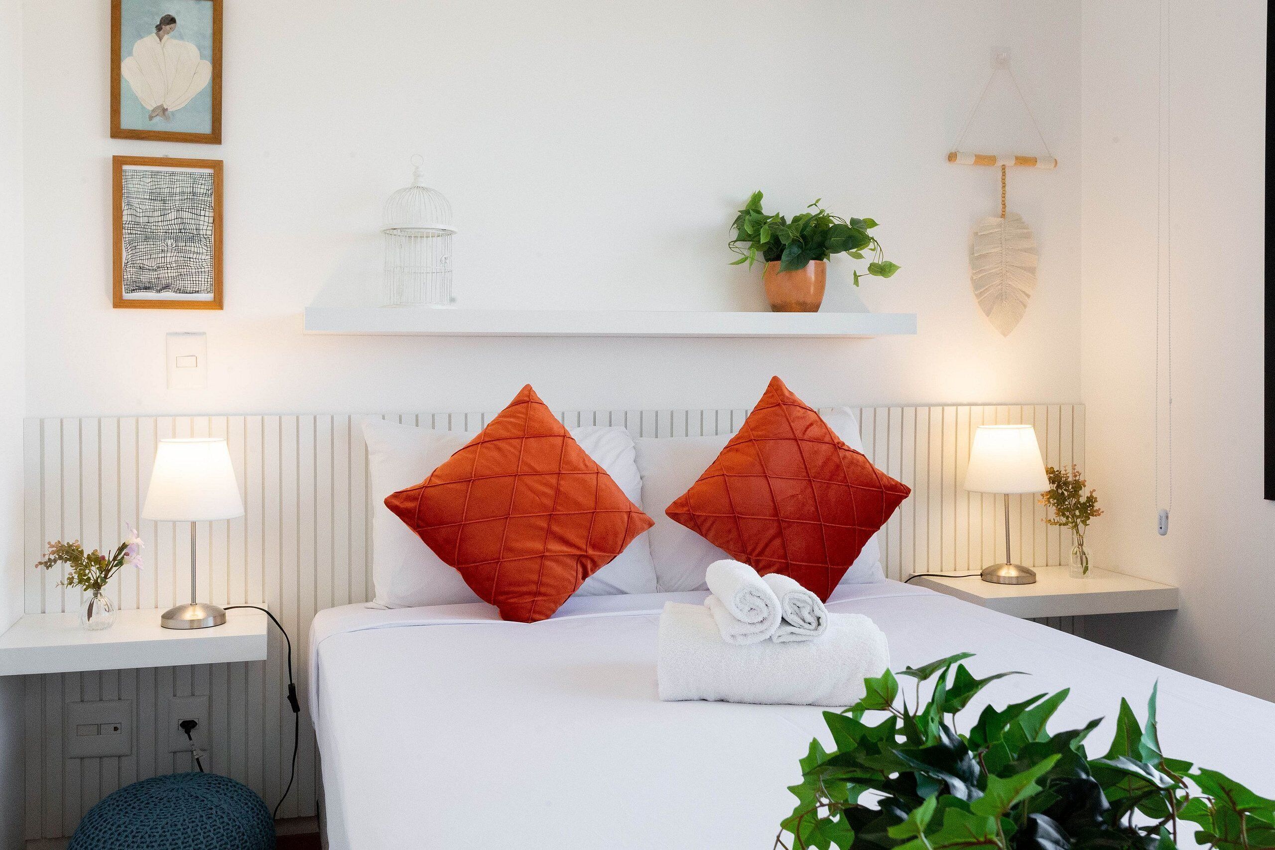 A neatly arranged hotel room with a bed, two nightstands, a shelf with decorative items, and a potted plant.