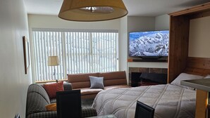 Une chambre d’hôtel moderne avec une table à manger en verre, un canapé, un lit, une télévision et une fenêtre avec des stores.