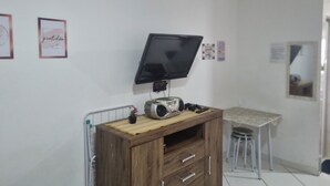 A room with a TV mounted on the wall, a wooden cabinet with drawers, a small table with a chair, and a radiator. - Kitnet na Cidade Ocian / Praia Grande (Praia Grande)