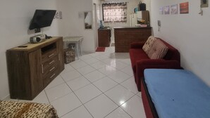 A room with a red sofa, a blue bed, a leopard-print chair, a wooden cabinet, a television, and a kitchen area in the background. - Kitnet na Cidade Ocian / Praia Grande (Praia Grande)