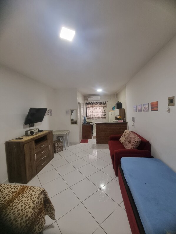 Quarto com sofá vermelho, cama azul, cadeira com estampa de leopardo, armário de madeira, televisão e área de cozinha ao fundo.