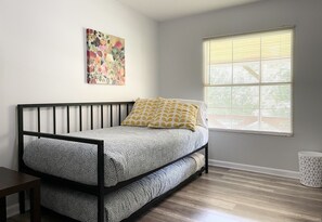 A room with a bed, a window with blinds, a wall-mounted painting, and a small trash can.