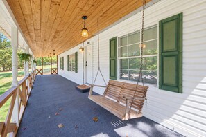 A covered porch with a wooden swing, white exterior walls, green shutters, and hanging lights.