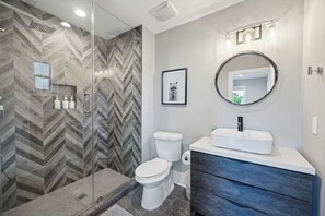 Un baño moderno con ducha de mosaico de espiga, inodoro blanco, tocador con espejo redondo y un cuadro en la pared.